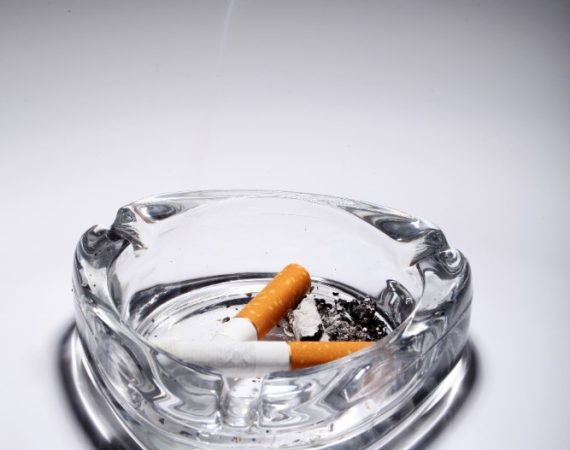 Smoke curls from a cigarette in a glass ashtray, accompanied by an assortment of rolling trays.